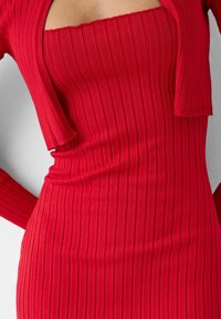 Czerwona sukienka z prążkowanej tkaniny z kwadratowym dekoltem i krótkimi rękawami, uszyta z elastycznego materiału. Tkanina ma pionowe linie, które nadają jej teksturę.