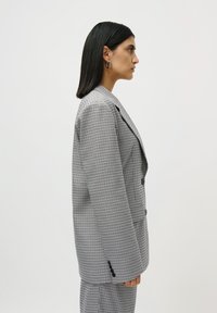Femeie îmbrăcată într-un costum cu carouri alb-negru, purtând cercei rotunzi din argint, stând în profil pe un fundal simplu, luminos.