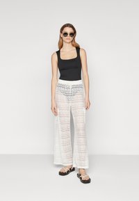 Débardeur noir ajusté, pantalon large en dentelle blanche transparente avec un motif en zigzag, et sandales plates noires. Accessoirisé avec des lunettes de soleil rondes.