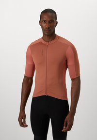 Maglia da ciclismo a maniche corte in un marrone rossastro chiaro, con zip frontale e pannelli texturizzati, abbinata a pantaloni ciclismo neri imbottiti.