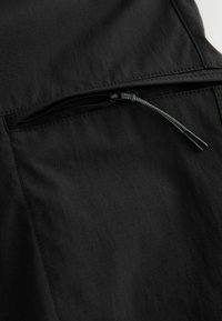 Schwarzer Stoff mit einer Reißverschlusstasche und kontrastierendem strukturiertem Muster. Der Reißverschluss verfügt über eine dünne, kordelartige Zuglasche für einfachen Zugriff.