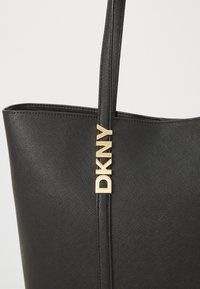 Musta olkalaukku, jossa on tekstuuri ja kultainen "DKNY"-logo. Varustettu tukevilla kahvoilla ja minimalistisella muotoilulla.