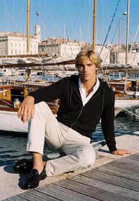 Jeune homme aux cheveux blonds portant une veste noire et un pantalon blanc, assis sur un quai près de voiliers dans une marina ensoleillée avec des bâtiments en arrière-plan.