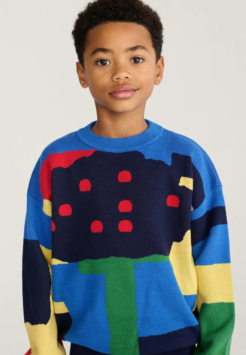 Garçon aux cheveux courts et bouclés portant un pull coloré avec des formes abstraites en bleu, vert, rouge, jaune et noir sur un fond uni.