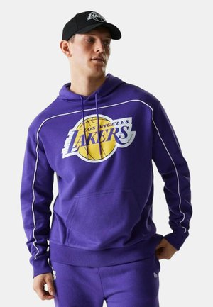 Uomo con felpa viola dei Los Angeles Lakers e pantaloni abbinati, con cappellino nero dei Lakers, in piedi davanti a uno sfondo semplice.