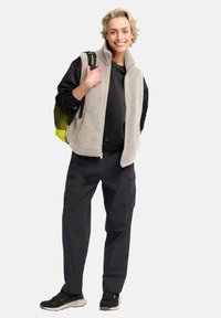 Gilet in pile grigio chiaro, camicia nera a maniche lunghe, pantaloni cargo grigio scuro e sneakers nere. Tiene uno zaino giallo.