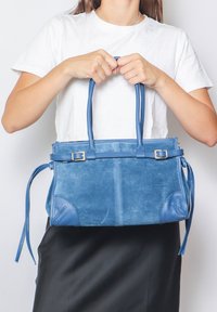 Borsa a mano blu in suede con dettagli in pelle, forma strutturata, manici doppi e legature laterali per una chiusura regolabile, posizionata di fronte a uno sfondo bianco.