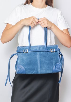 Borsa a mano blu in suede con dettagli in pelle, forma strutturata, manici doppi e legature laterali per una chiusura regolabile, posizionata di fronte a uno sfondo bianco.