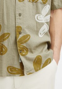 Camisa de manga corta de tela beige con grandes estampados florales en marrón y blanco. Presenta botones, un bolsillo y un dobladillo ligeramente curvado.