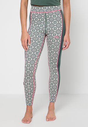 Frau trägt grüne und weiß gemusterte Leggings mit pinkfarbenen Paspeln und einfarbigen grünen Seitenpaneelen, steht barfuß vor einem weißen Hintergrund.