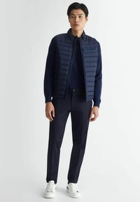 Gilet imbottito blu navy con design a cuciture verticali, indossato sopra un maglione coordinato e pantaloni scuri, abbinato a sneakers bianche.