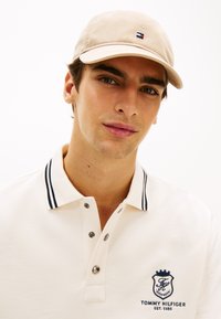 Καπέλο ανοιχτό μπεζ με μικρό λογότυπο, σε συνδυασμό με λευκό πόλο πουκάμισο με σκούρο ριγέ γιακά και κεντημένο λογότυπο στο στήθος.