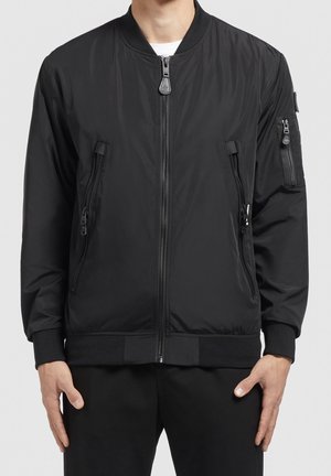 Mężczyzna ubrany w czarną kurtkę bomberkę z zamkiem błyskawicznym z przodu oraz kieszeniami na piersi i na rękawie, w połączeniu z czarnymi spodniami.