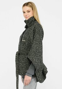 Poncho a pattern bianco e nero con colletto alto, maniche larghe, cintura e logo ricamato. Tessuto a maglia testurizzato, vestibilità rilassata.