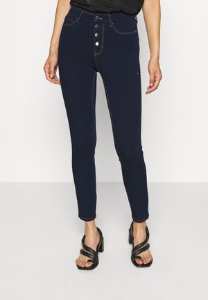 Vrouw draagt donkerblauwe high-waisted skinny jeans met knoopsluiting en zwarte sandalen met hak, tegen een witte achtergrond.