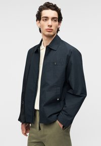 Veste légère bleu marine avec col, boutons sur le devant et une seule poche poitrine. Tissu texturé et coupe décontractée. Associée à un pantalon olive.