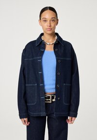 Donkerblauwe spijkerjas met contrasterende stiknaden, knoopsluiting en twee voorkantzakken. Gedragen over een lichtblauwe trui.