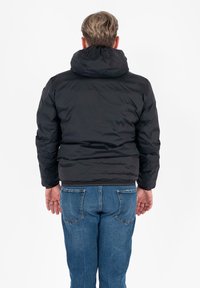 Zwarte pufferjack met een hoge kraag en capuchon, met een gewatteerd ontwerp. Draagt het met blauwe jeans, waarbij de achterkant zichtbaar is.