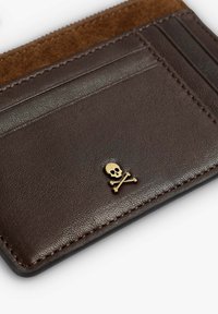 Tarjetero de cuero marrón con un detalle de ante, múltiples ranuras para tarjetas y un emblema dorado de calavera con huesos cruzados. Textura suave, diseño compacto.