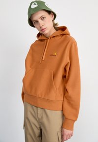 Orange hoodie made of soft fabric featuring a front pocket and a small yellow logo. Paired with light tan cargo pants and a green bucket hat.