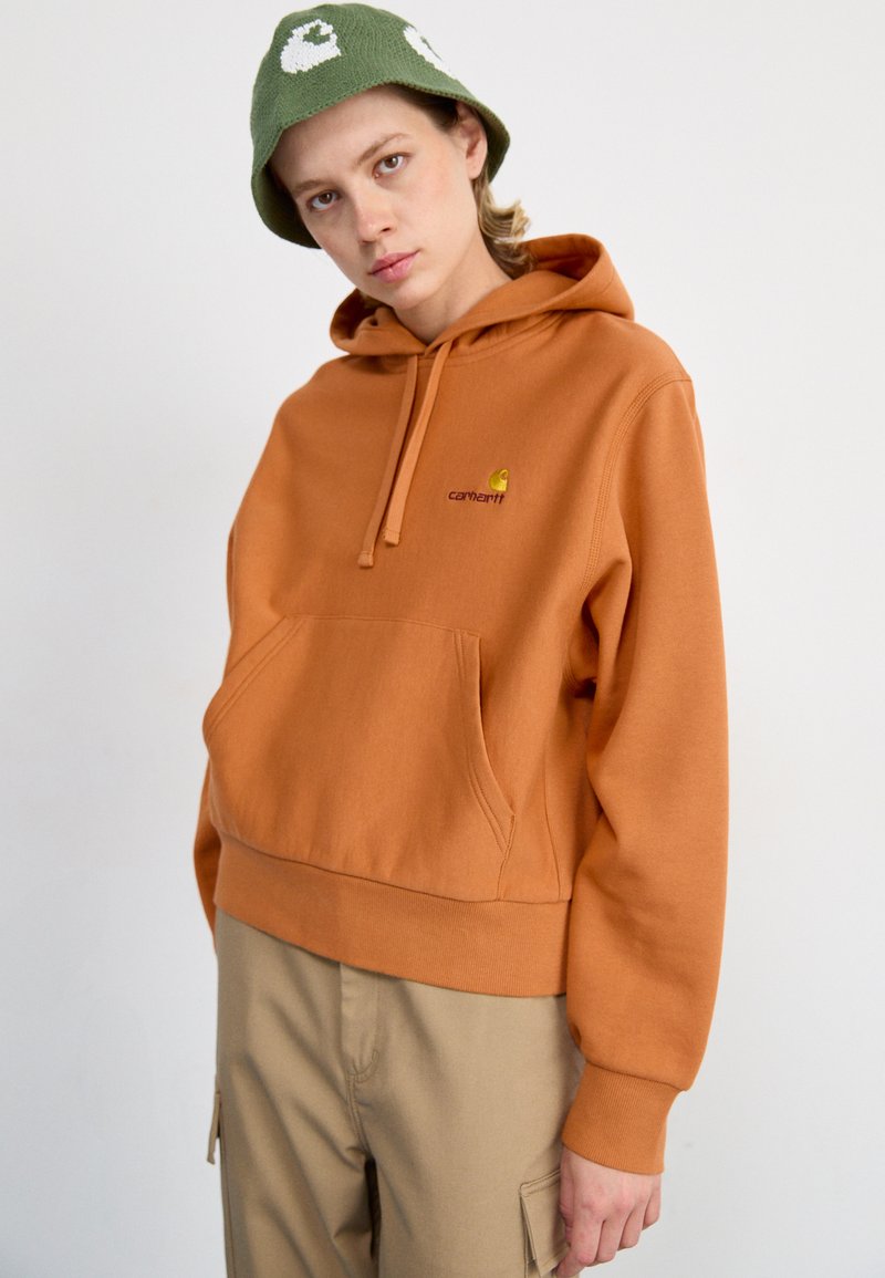Orange hoodie made of soft fabric featuring a front pocket and a small yellow logo. Paired with light tan cargo pants and a green bucket hat.