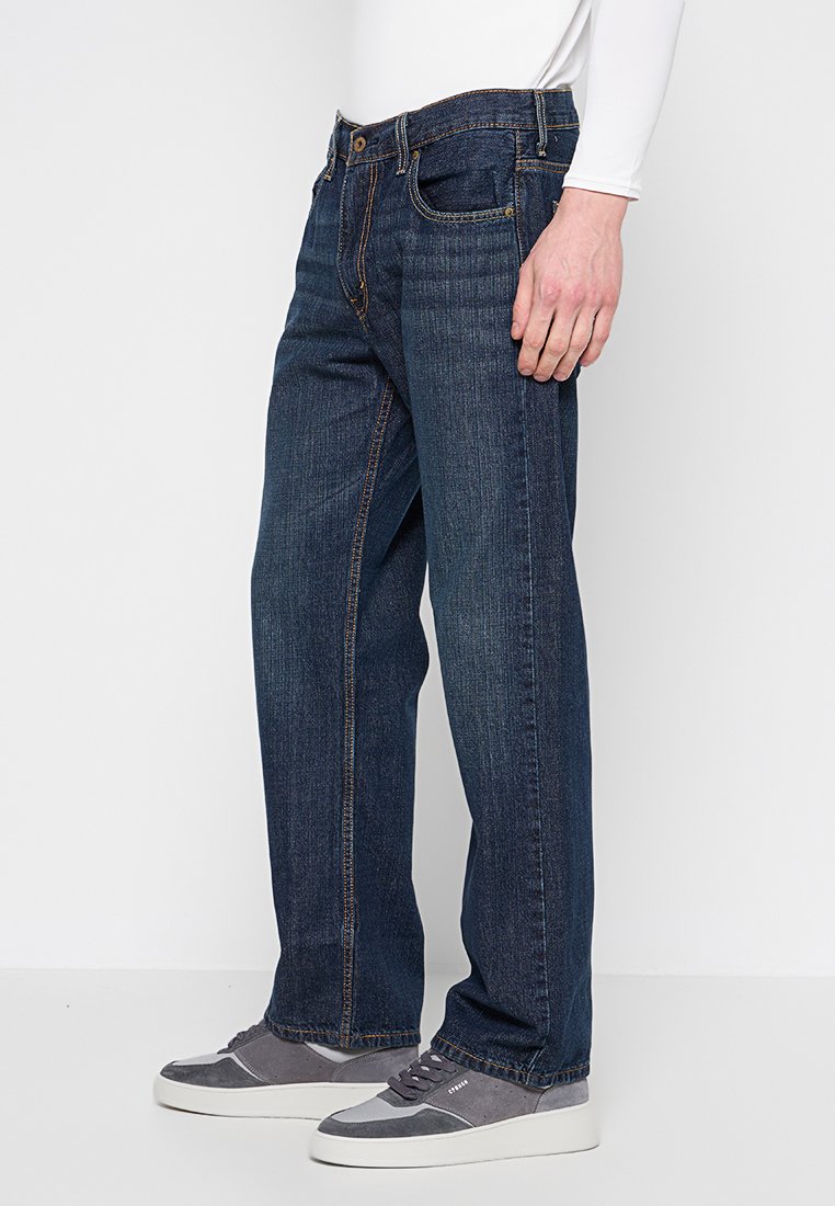 Jean en denim bleu foncé avec une coupe droite, cinq poches et des coutures orange contrastantes, portés avec des baskets grises.