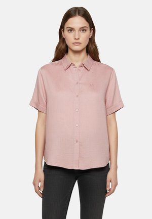 Donna dai capelli castano lunghi indossa una camicia rosa chiaro a maniche corte con bottoni e pantaloni neri, in piedi davanti a uno sfondo semplice.