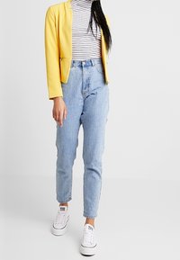 Blazer jaune porté sur un col roulé rayé, associé à un jean taille haute bleu clair et des baskets blanches. Tissu lisse et design ajusté.
