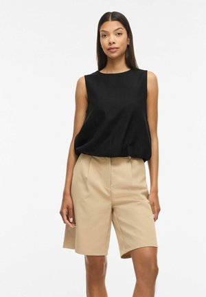 Kvinde iført en ærmeløs sort top og beige knælange skræddersyede shorts, stående mod en ensfarvet hvid baggrund.
