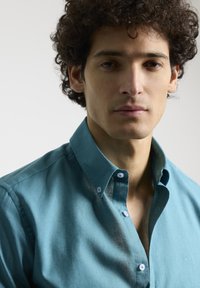 Camisa de botones turquesa con un cuello estructurado, textura suave y un ligero brillo. Cuenta con botones blancos y un pequeño detalle de logo.