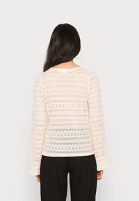 Top en dentelle à manches longues de couleur beige clair, avec un motif floral rayé et des manches évasées, associé à un pantalon noir à jambes larges.