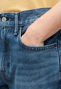 Main droite insérée dans la poche avant d'un jean bleu porté avec un haut jaune.