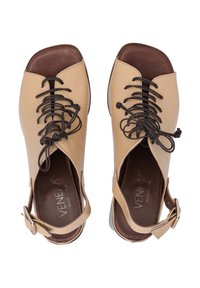 Beige Leder Sandalen mit offenen Zehen, die dunkle Schnürsenkel, ausgeschnittene Seiten und eine braune Innensohle aufweisen. Verstellbarer Knöchelriemen mit Beschlägen in Goldoptik.