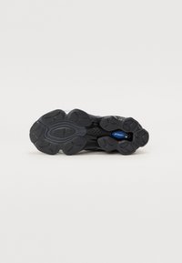 Semelle de chaussure de sport noire avec un motif de bande de roulement texturé et un petit détail ovale bleu dans la zone de la voûte plantaire, isolée sur fond blanc.