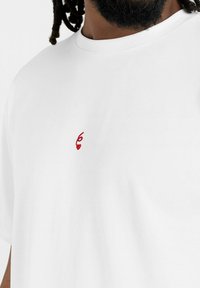 Vit kortärmad t-shirt i bomull, med ett litet rött broderat nummer '6' och ett hjärtmönster på vänstra bröstområdet.