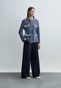 Nő, aki kék, texturált, öves dzsekit visel, fehér szegélyű zsebekkel, és széles szárú, sötét nadrágot, egyik kezét a zsebében tartva áll.