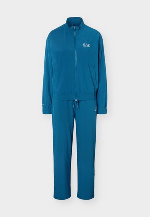Personne debout portant un survêtement bleu EA7 avec une veste zippée et un pantalon assorti, associé à des baskets claires sur fond blanc.