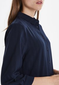 Camicia blu navy con colletto, caratterizzata da righe verticali, chiusura con bottoni e maniche lunghe. Il tessuto appare liscio e leggero.