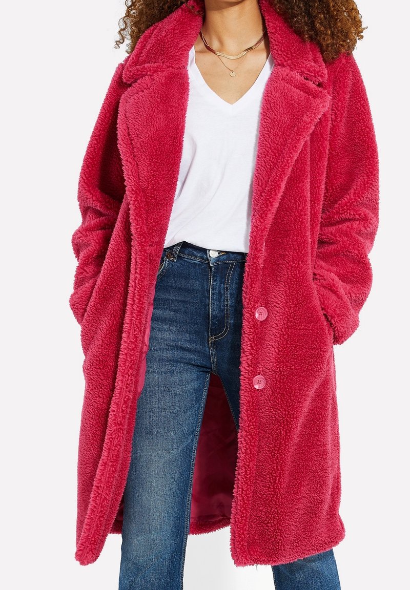 Femme portant un long manteau teddy rose vif par-dessus un t-shirt blanc à col en V et un jean bleu, les mains dans les poches du manteau.