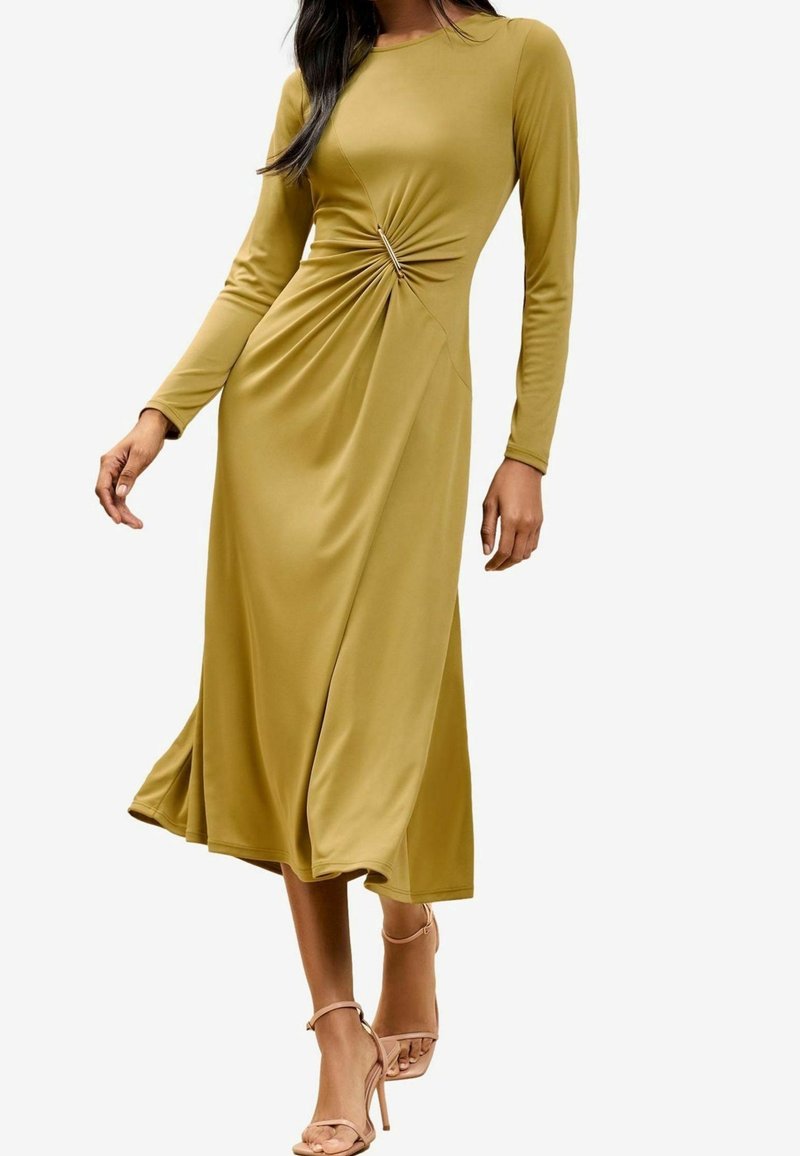 Femme portant une robe midi jaune moutarde à manches longues avec un détail froncé sur le côté, associée à des sandales à talons hauts nude à brides.