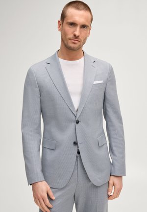 Man draagt lichtgrijs tweeknops pak over wit T-shirt, staat voor een effen lichte achtergrond, handen ontspannen langs het lichaam.