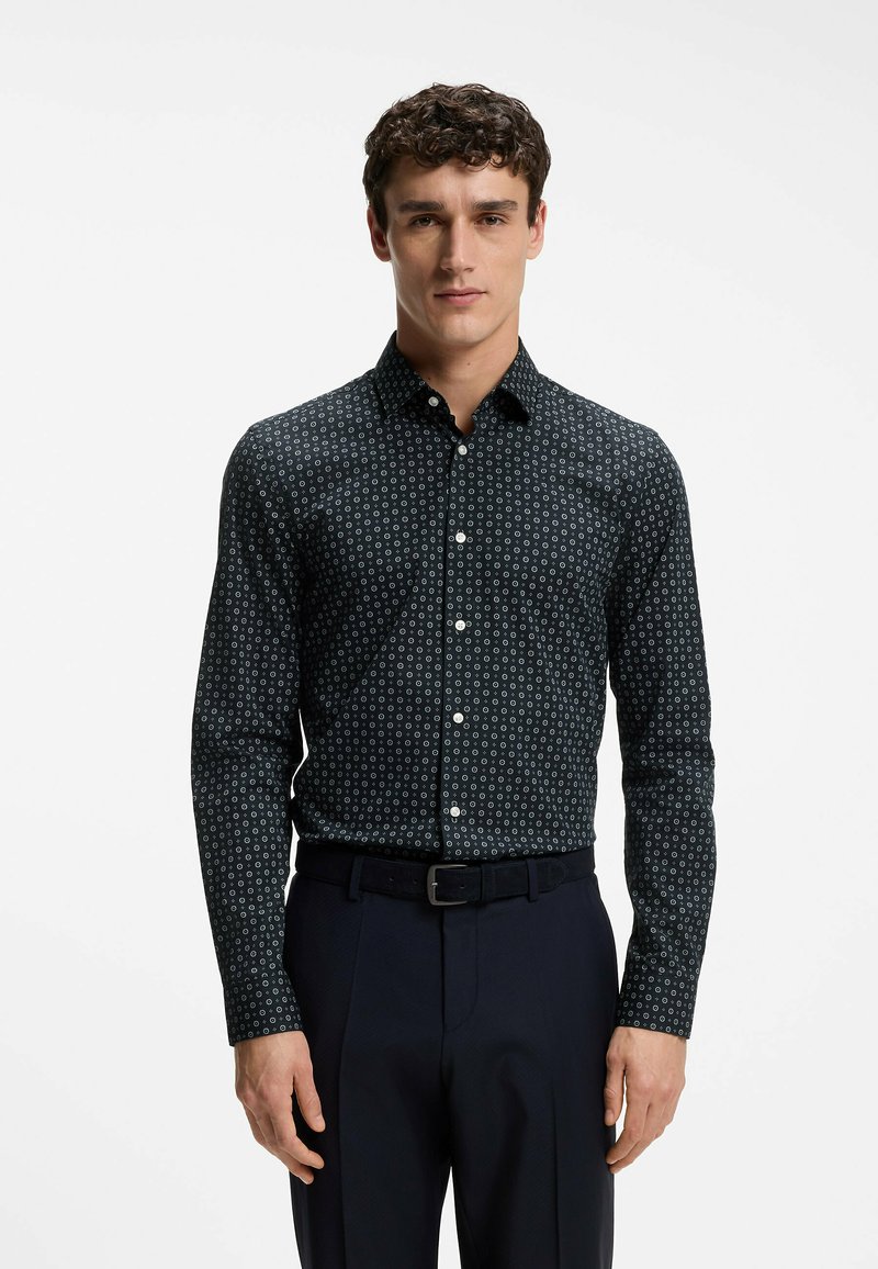 Homme portant une chemise noire à boutons avec de petits motifs circulaires blancs, rentrée dans un pantalon foncé avec une ceinture noire, debout devant un fond blanc.