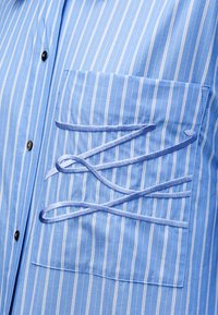 Ανοιχτό μπλε ριγέ πουκάμισο με τσέπη στο στήθος· διαθέτει διακοσμητικές μπλε θηλιές από ύφασμα στην τσέπη. Το ύφασμα φαίνεται λείο και ελαφρώς γυαλιστερό.