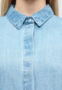Jasnoniebieska koszula jeansowa z ostrym kołnierzykiem, zapięciem na guziki oraz pionowymi przeszyciami. Materiał wykazuje subtelną fakturę i wzór prania.