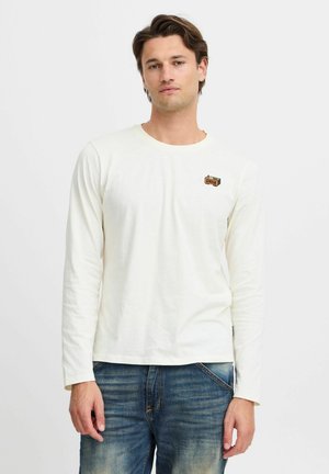 Long-sleeve ivory t-shirt in cotton, featuring a chest graphic in red and green, with a rounded neckline and standard fit. Denim jeans worn.