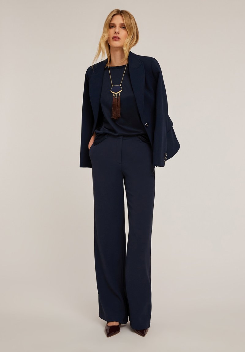 Abito blu navy su misura con giacca aderente e pantaloni a gamba larga, abbinato a un top scuro e a una lunga collana decorativa con frange.