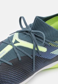 Scarpa sportiva con una tomaia in mesh testurizzato di un verde scuro con accenti giallo neon e una base nera con un motivo lineare sfumato.