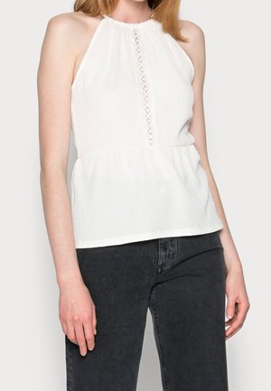 Frau trägt ein ärmelloses weißes Neckholder-Top mit Spitzen-Detail in der Mitte, kombiniert mit hoch taillierten schwarzen Jeans.