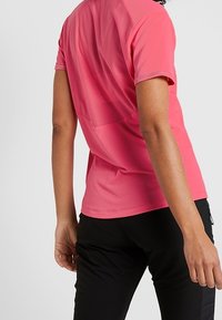 Short-sleeve athletic shirt in bright pink, featuring a textured back panel and a smooth front. Paired with black pants, showing a fitted design.