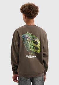 Bruine crewneck sweatshirt van zacht materiaal, met een groot grafisch ontwerp van een "B" met groene bladeren eroverheen en logo-tekst eronder.